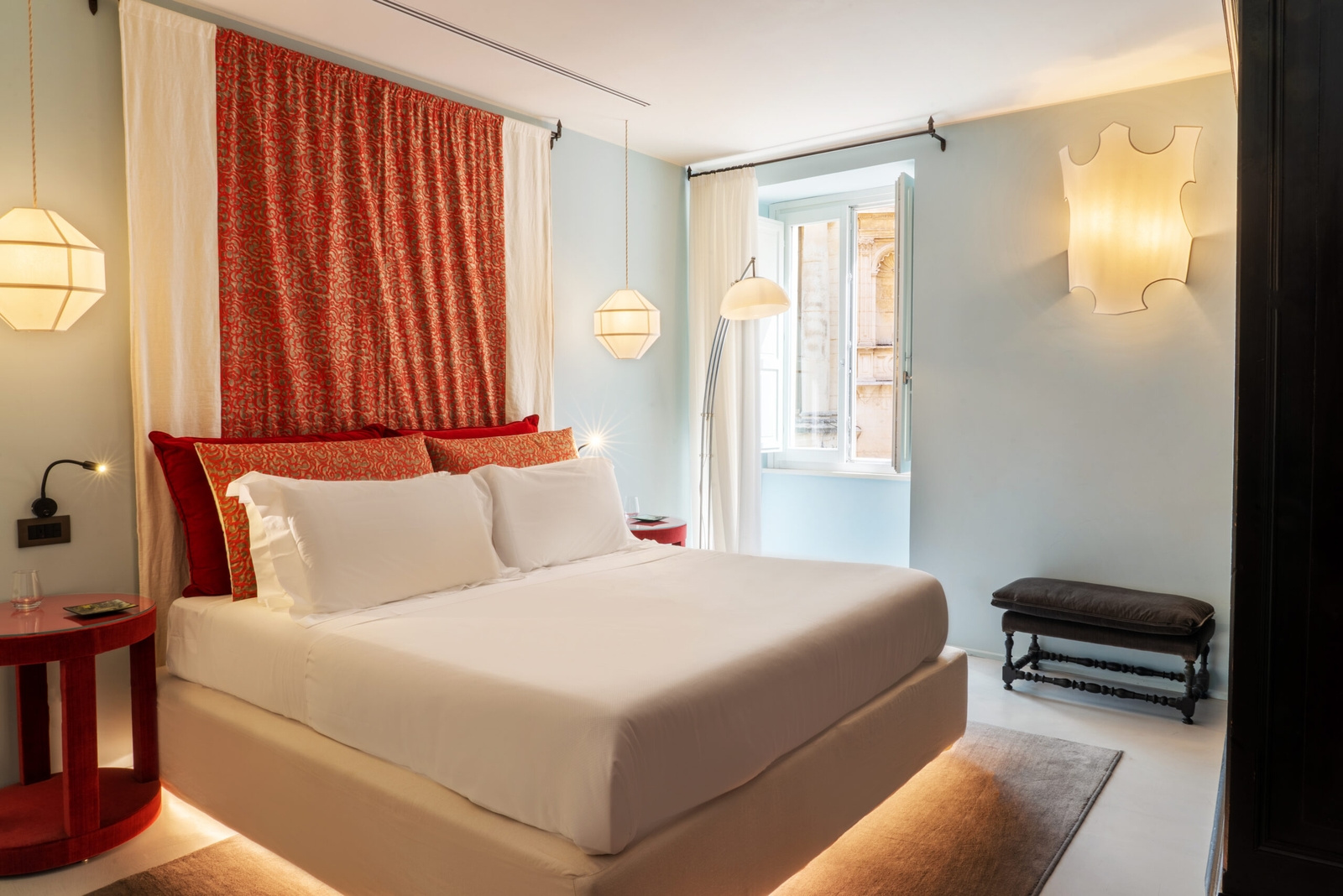 Interior of a bedroom with a double bed with white sheets and behind it an orange curtain and headboard. The walls are light blue and there is an open window.