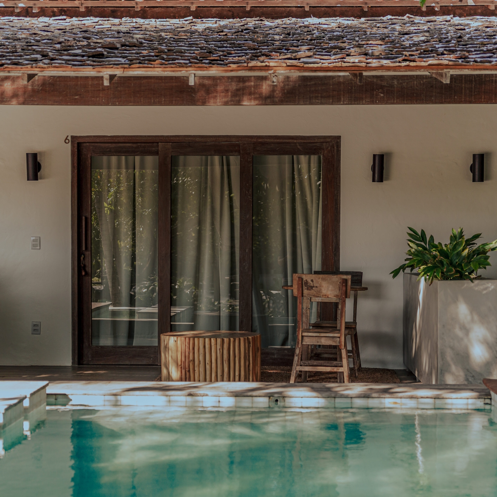 Exterior of a terrace and doorway into a bedroom. In the foreground is a blue pool with wooden chairs next to it.