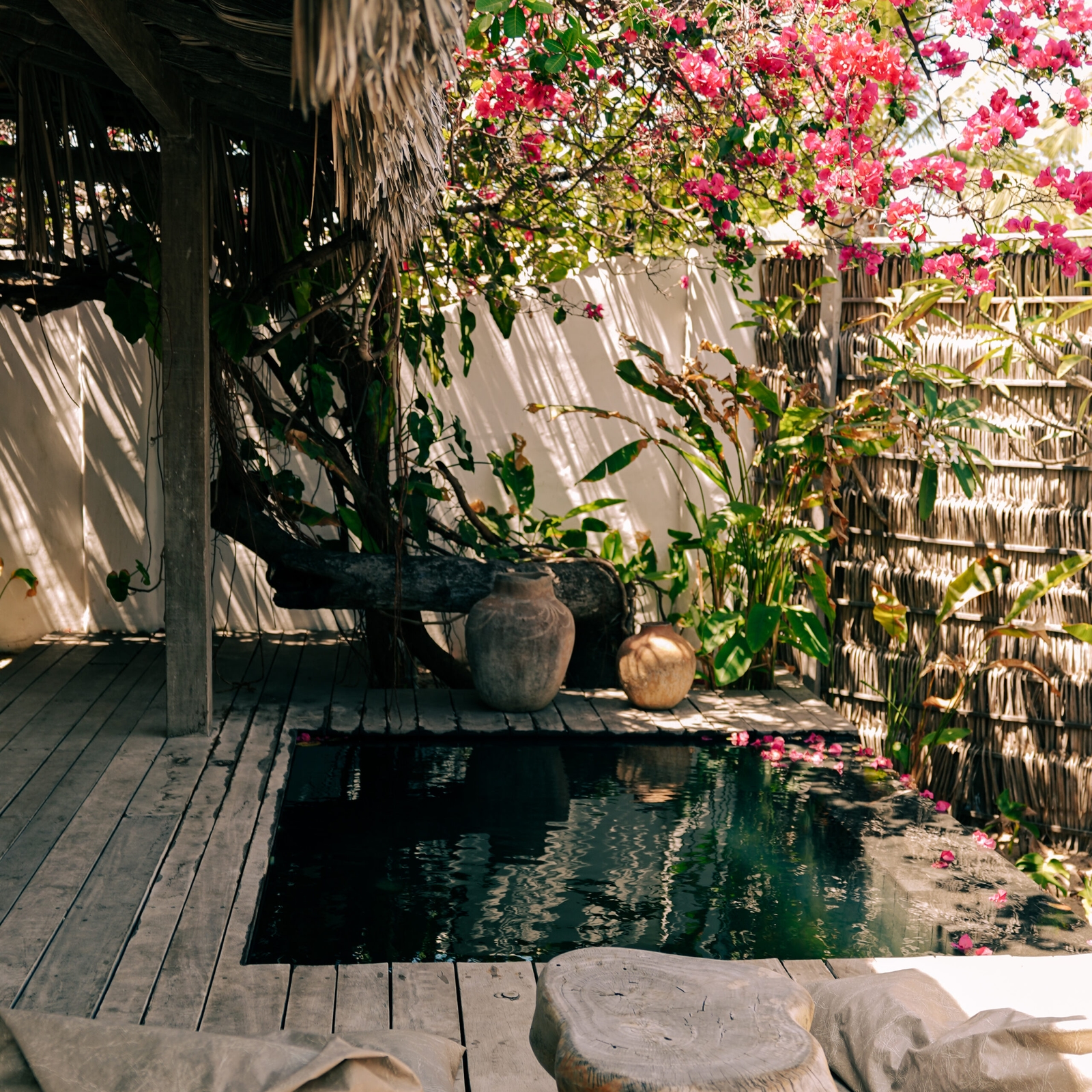 Exterior of a soaking tub surrounded by wooden decking, a thatched sunshade terrace and pink flowers, vines and ferns.