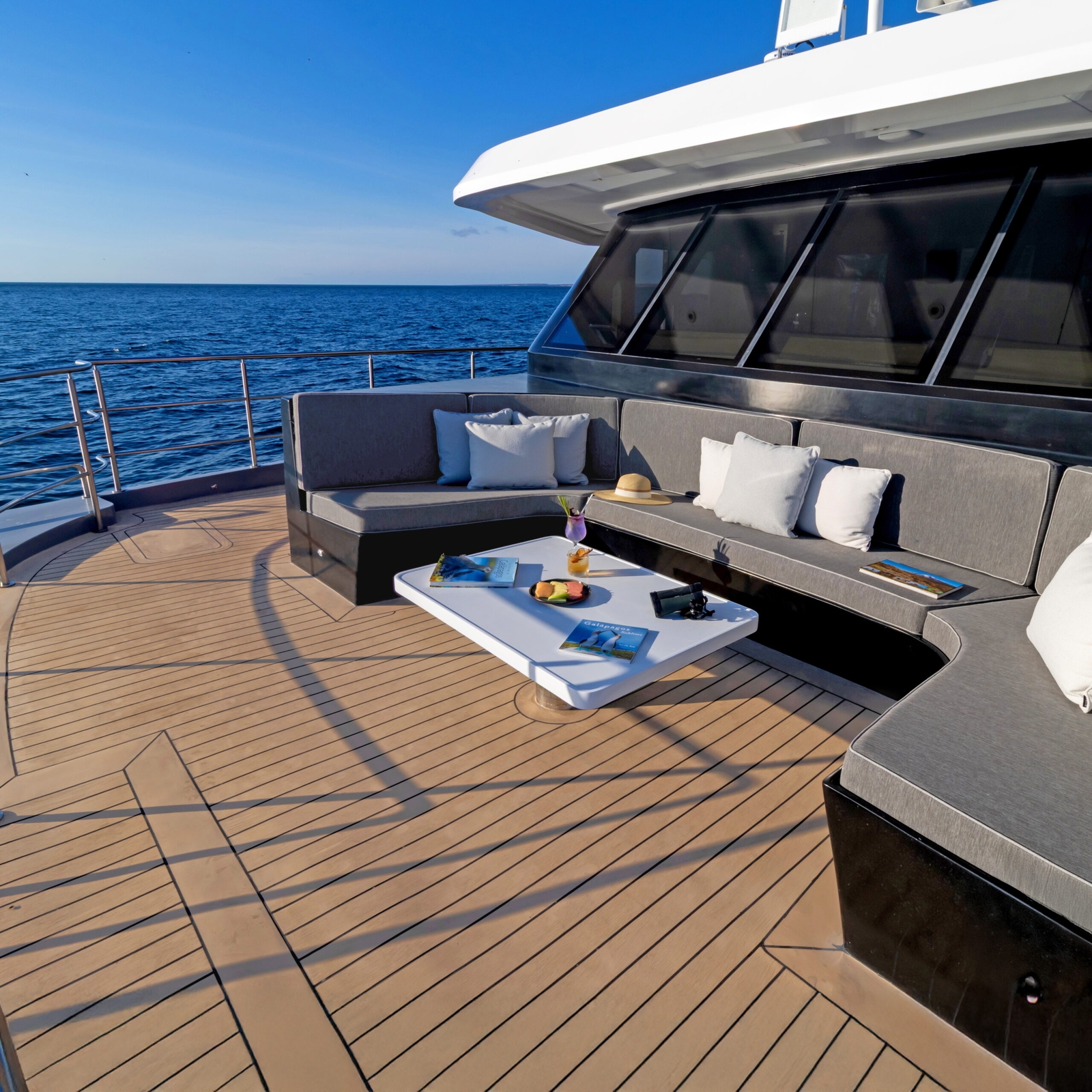 Open ship deck with view of the front windows of the ship, a banquette with seating and snacks, and open sea in the background.