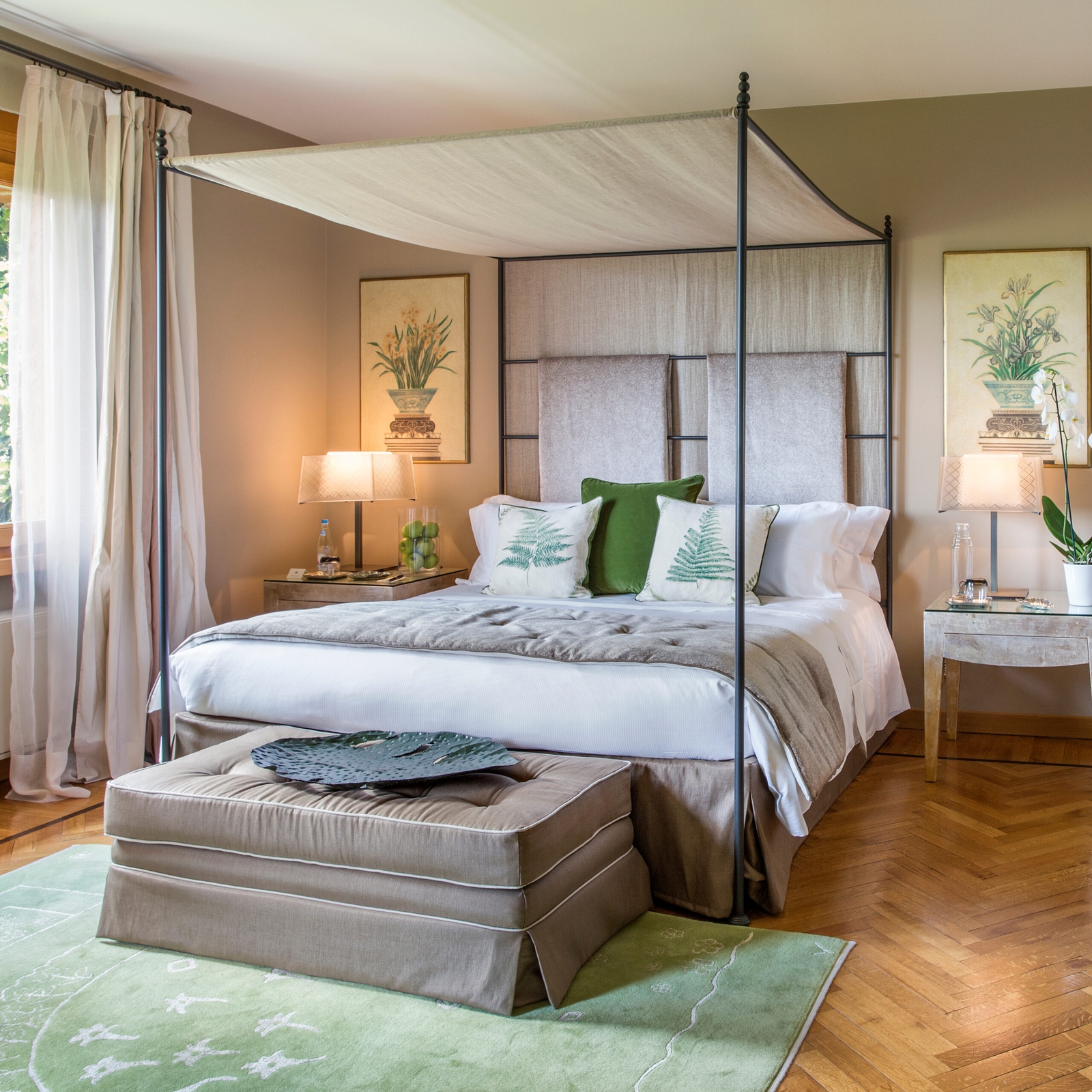 Bedroom scene showing a double bed with white and green and grey sheets, and a green plush rug, warm lighting, two bedside lamps and a canopy over the bed.