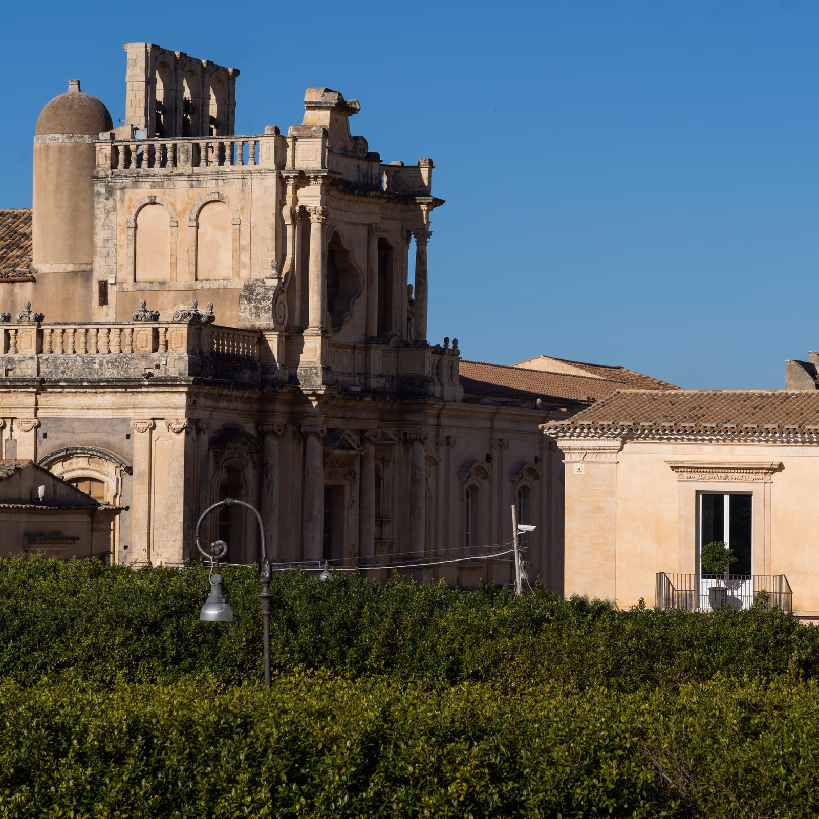Exteriors of historic Noto buildings with green hedges in the foreground.