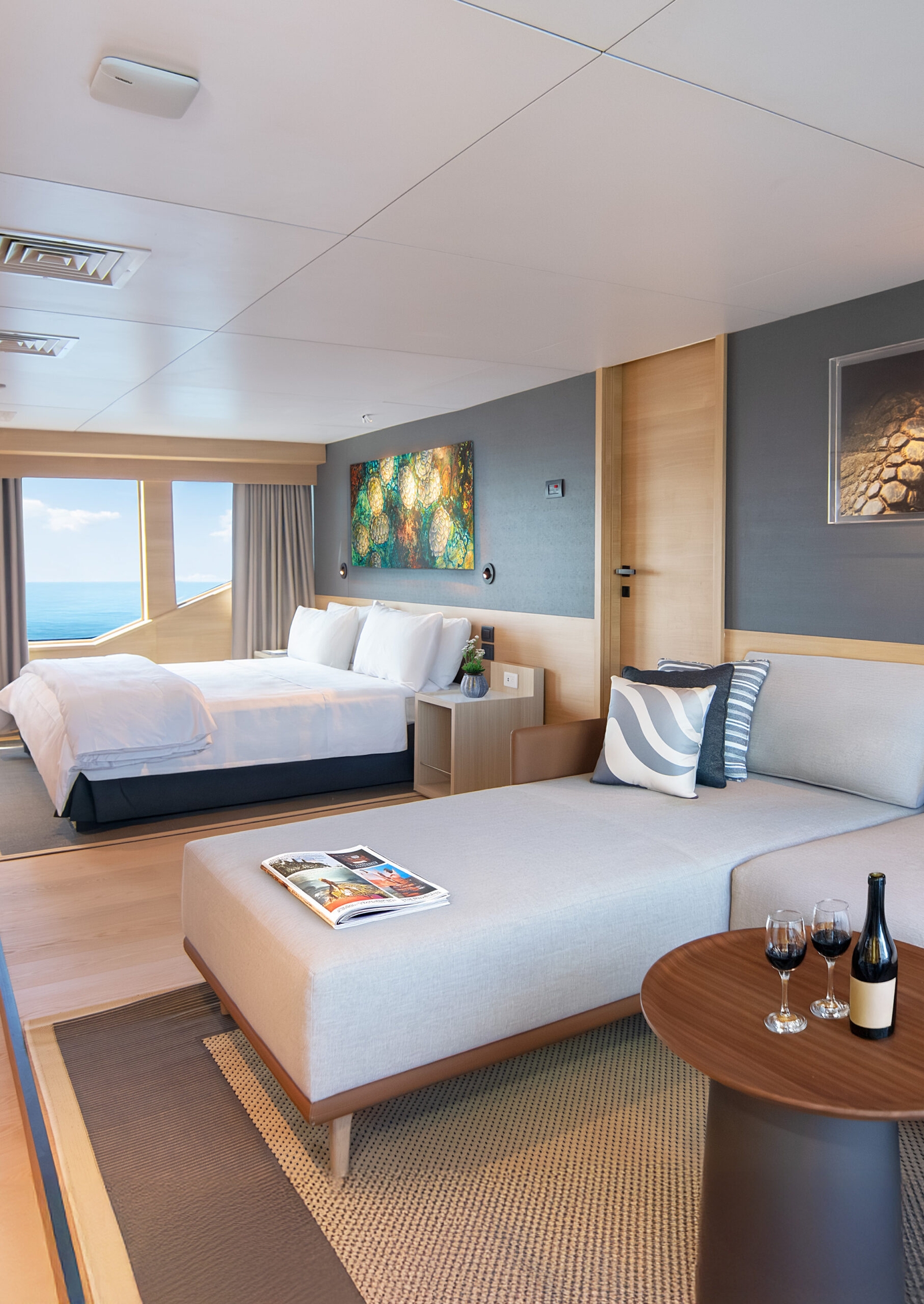 Bedroom with a chaise lounge in the foreground with pillows and magazines and a bottle of wine with two glasses, and in the background a double bed and window out onto the ocean.