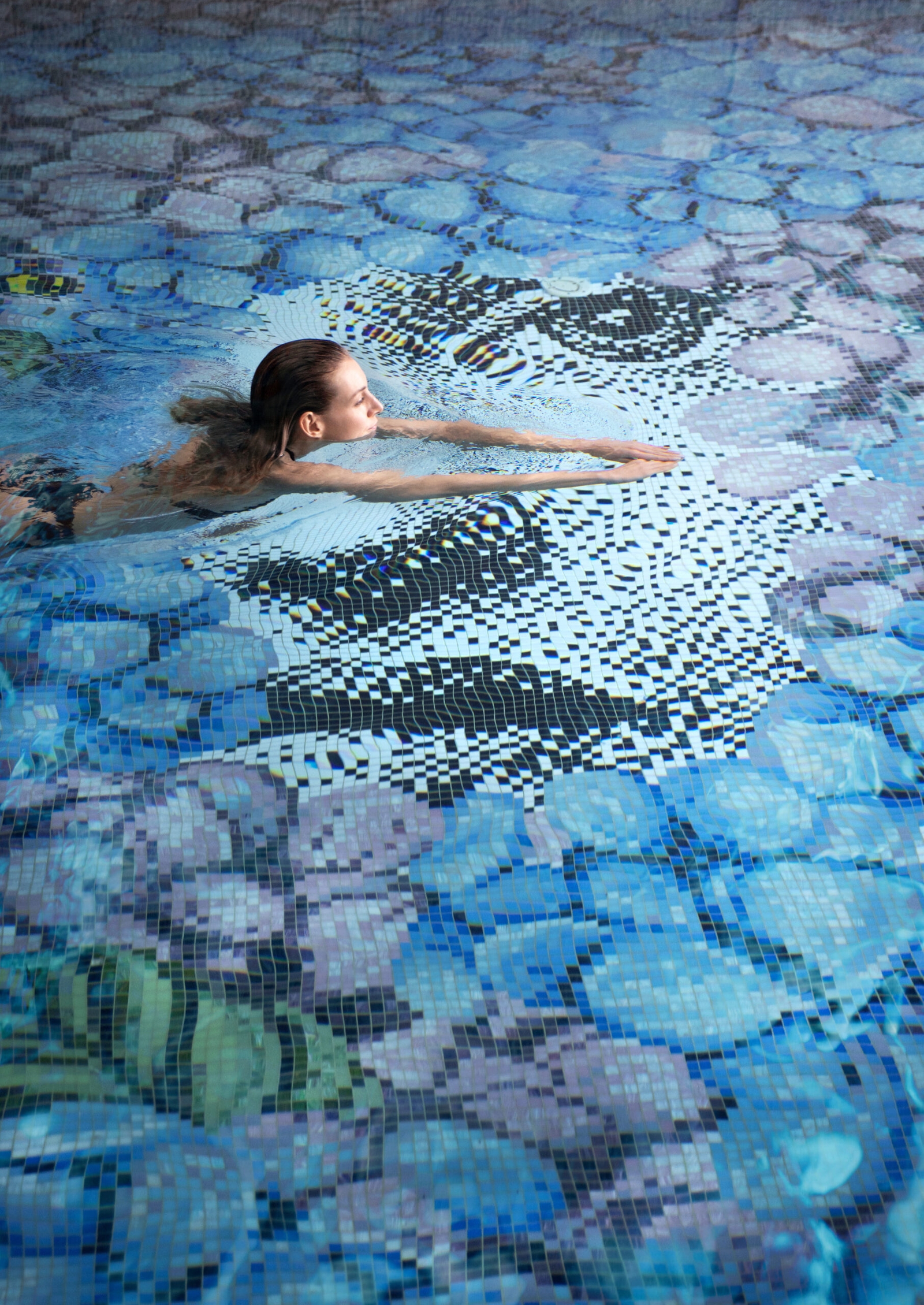Woman swimming in a blue pool with a mosaic floor.