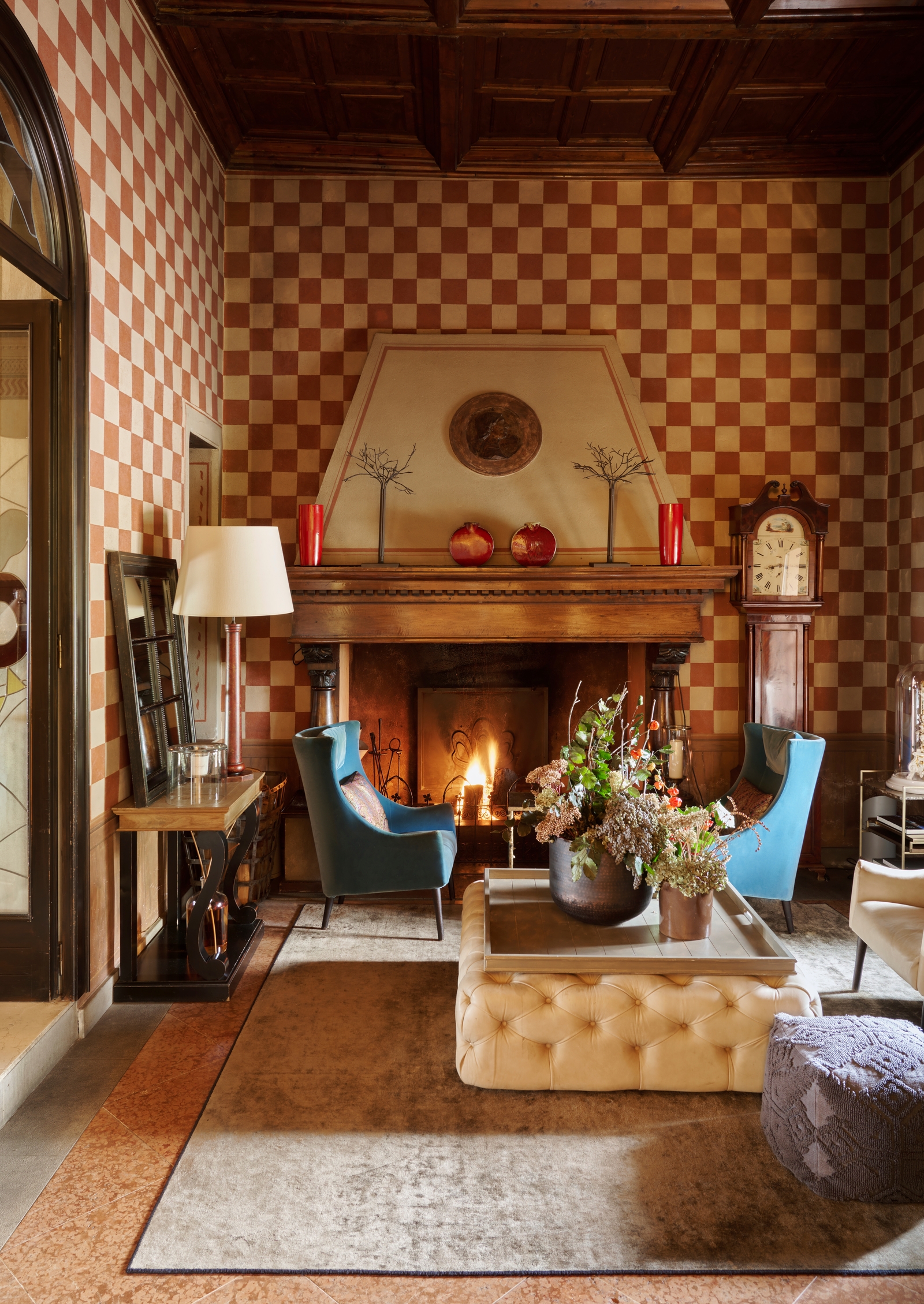 Interior scene of a living area with two blue plush chairs by a fireplace, rugs, coffee tables, lamps and red and white checked wallpaper.