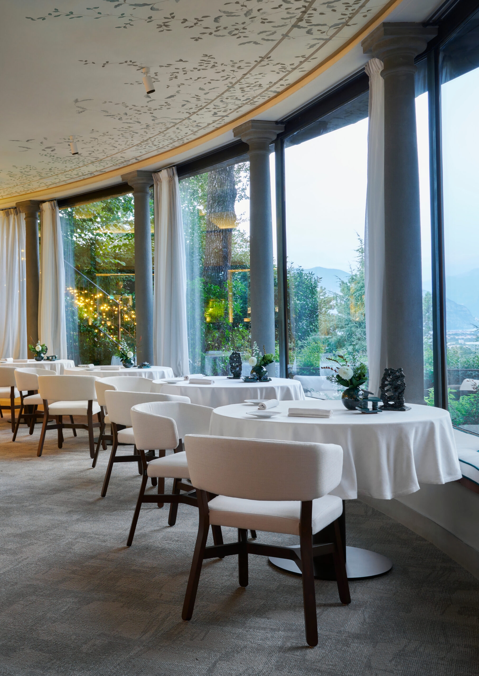Several formal tables dressed in white tablecloths with white dining chairs in a grey carpeted room with large floor-to-ceiling picture windows overlooking a nature scene.