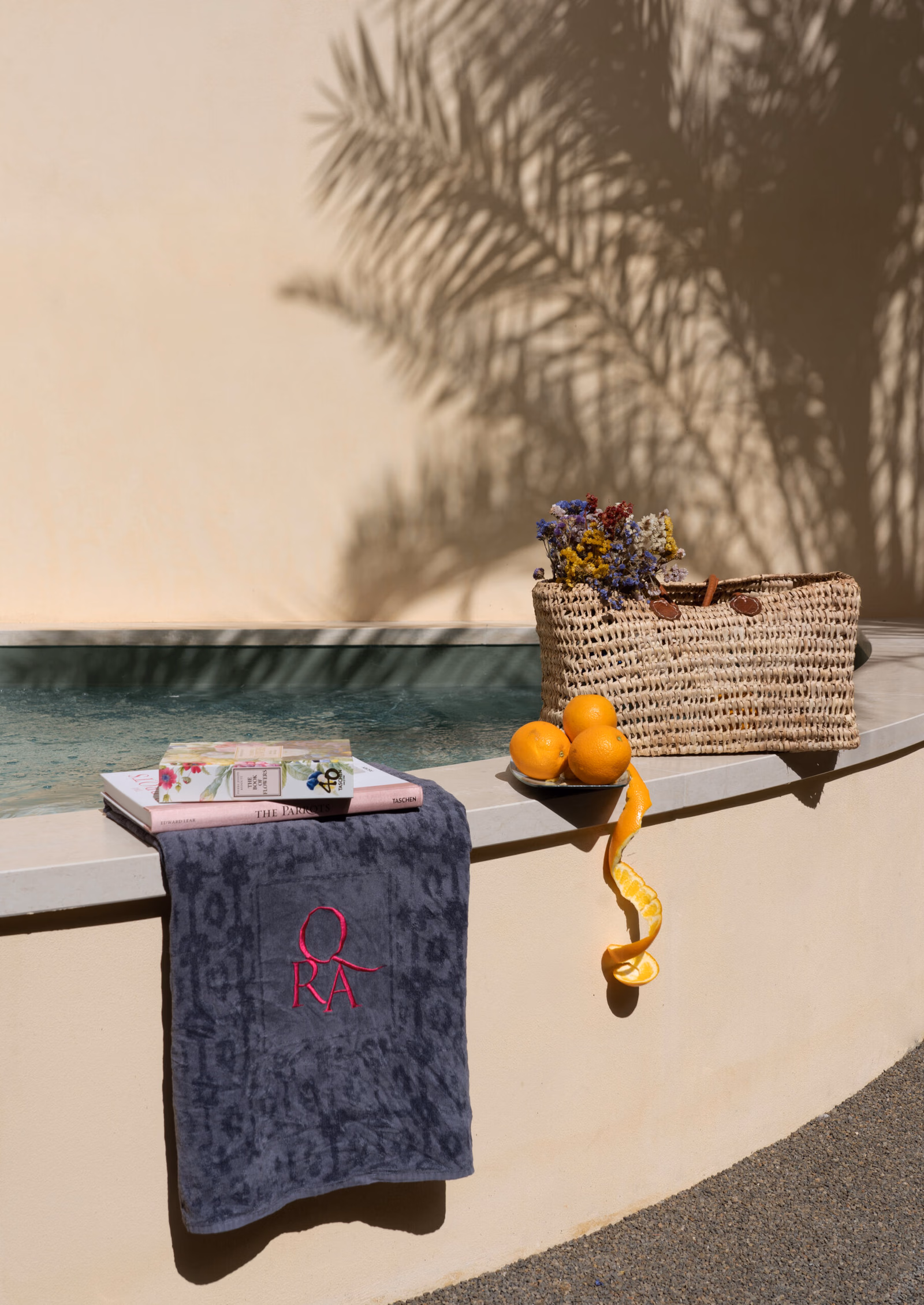 Exterior of the ledge of a pool with a towel, orranges and snacks, and the shadow of a palm tree.