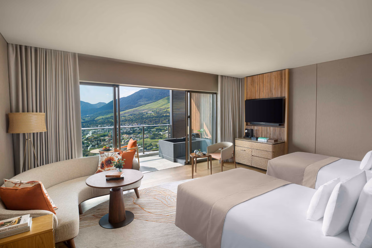 A bedroom with beige and white decor, two beds, a sofa and a view of the mountains