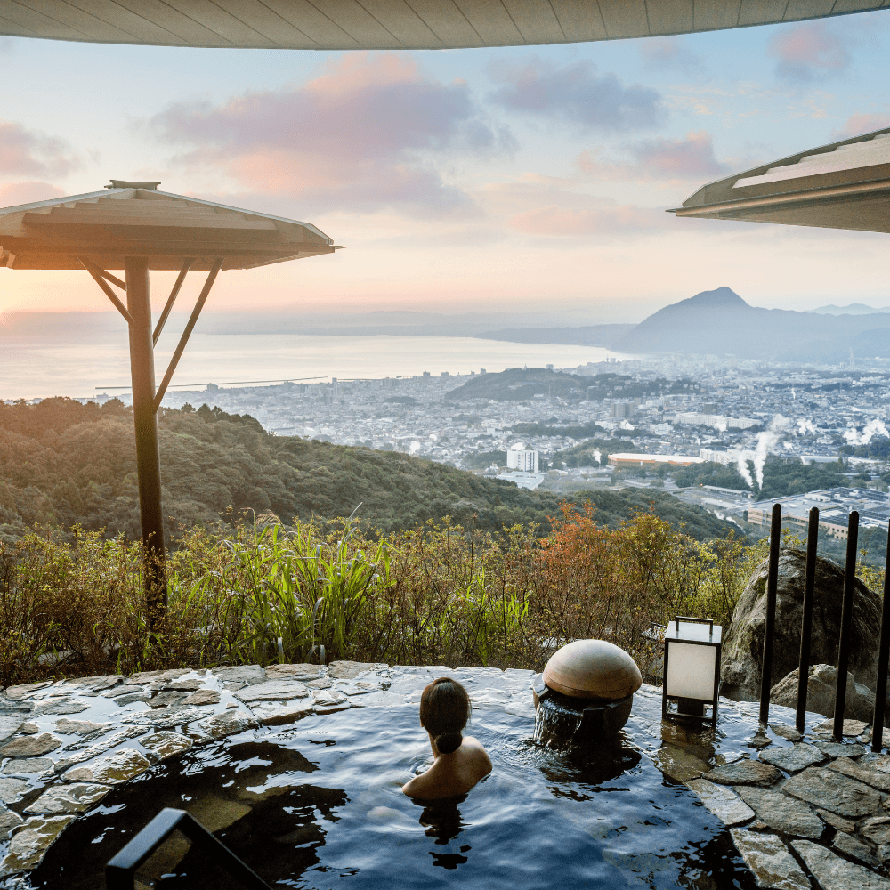 A woman in an outdoor onsen looking out over a sunset view of the mountains and sea