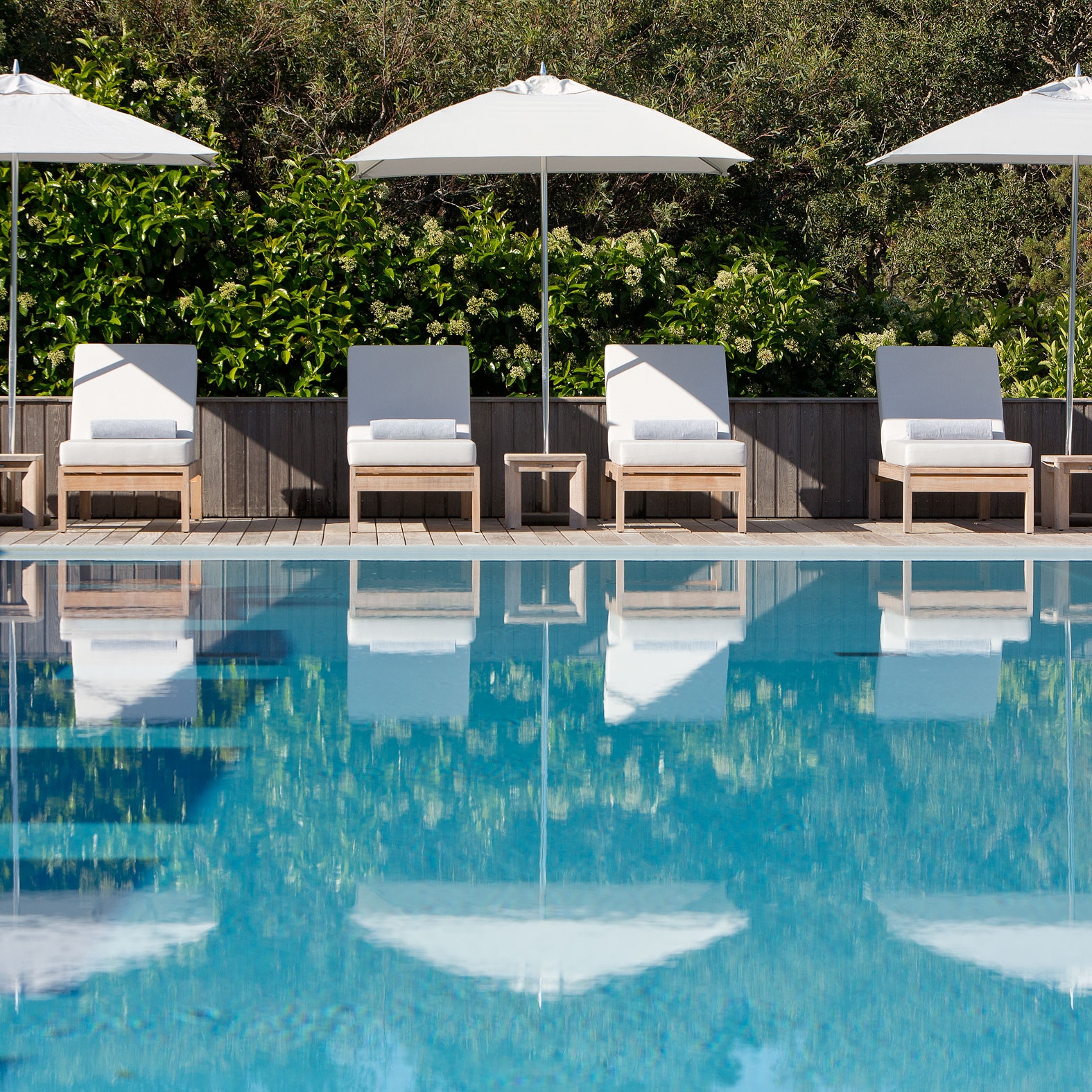 Blue pool water in foreground with four white sun loungers and umbrellas in the background.