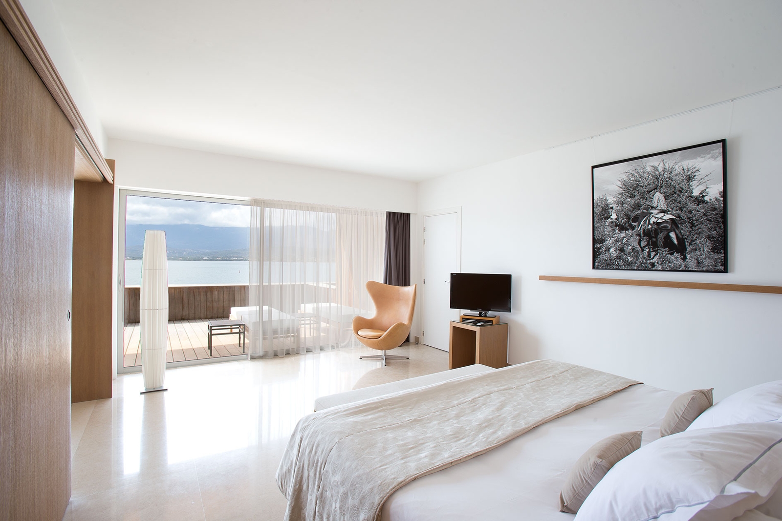 Interior shot of a white hotel room with white bed in the foreground, black and white picture on the wall and the room opens onto a deck overlooking the sea.