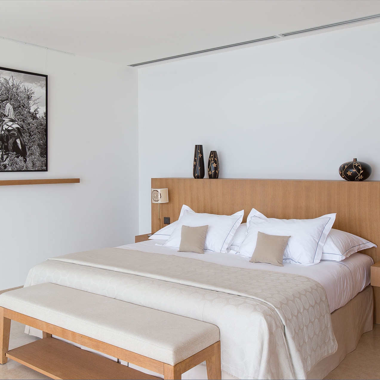 Interior shot of a bedroom with a blonde wood bed and white bedding plus a black and white photo on the wall.