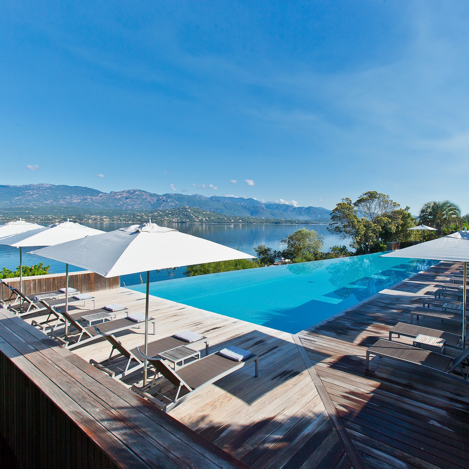 Pool with blue water, sun loungers and umbrellas along a wooden deck, and in the background it overlooks the sea.