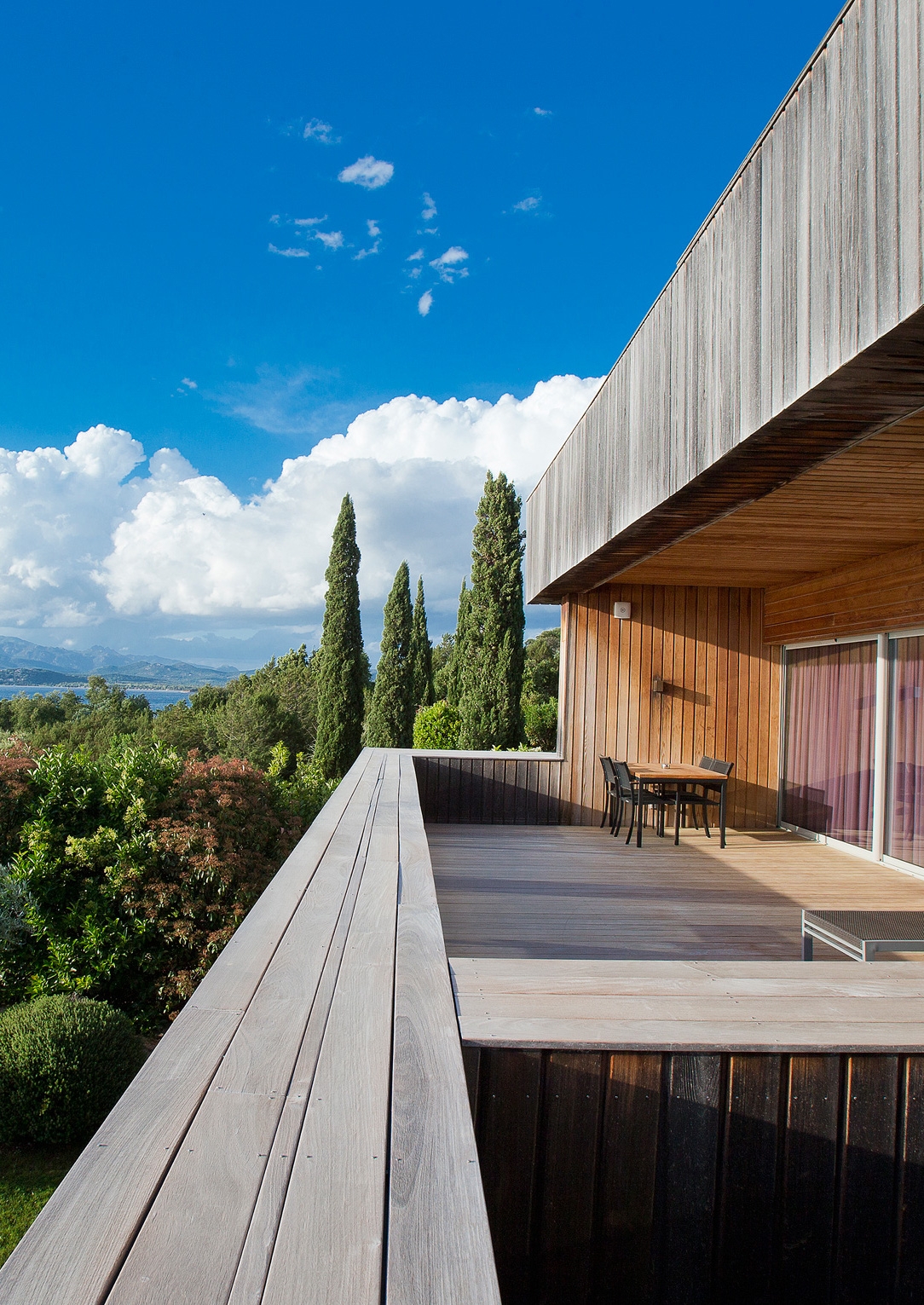 Exterior of a row of room decks with natural wood walls and outside are tall evergreen trees, clouds, blue sky and greenery beyond.