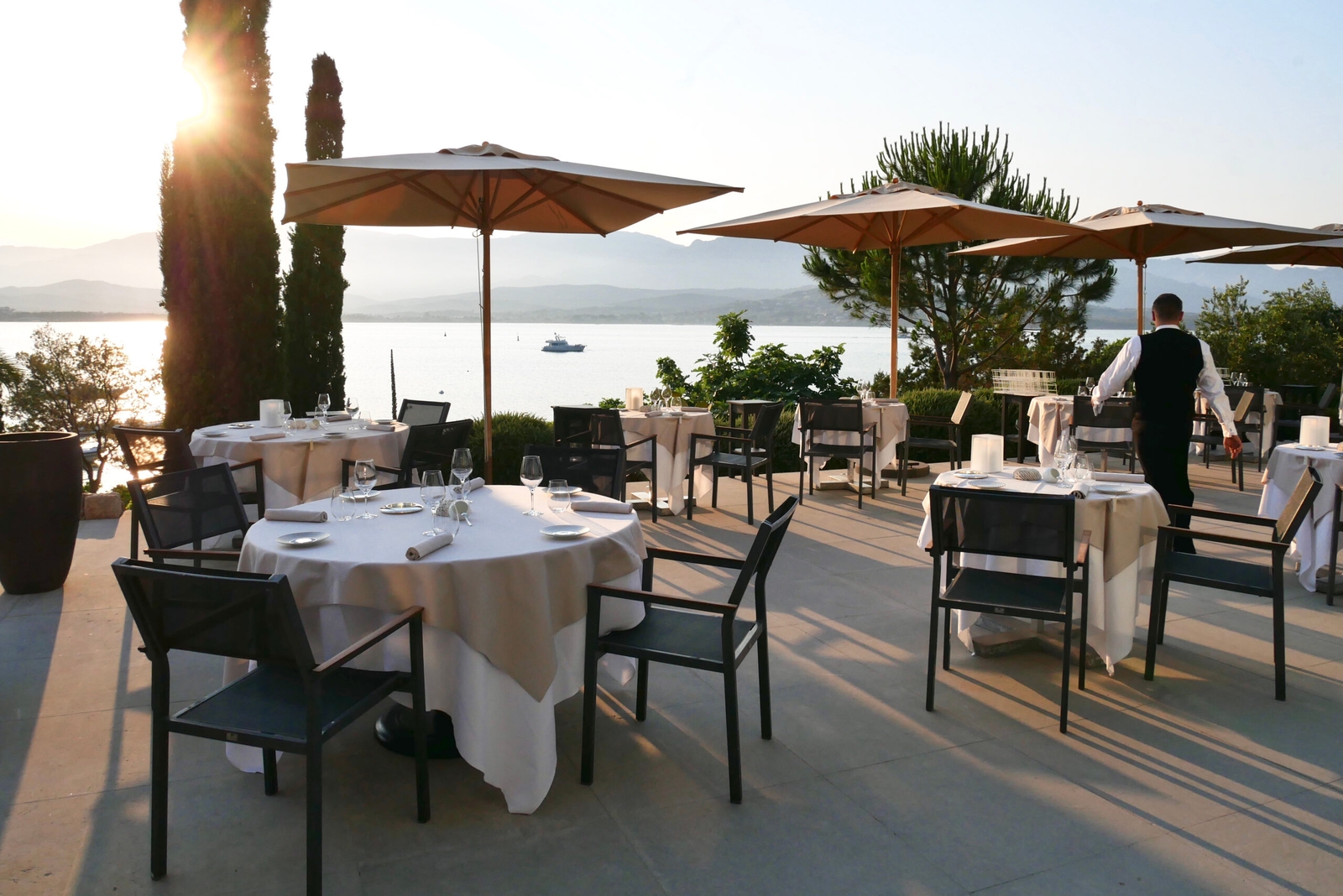 Tables with white tablecloths and umbrellas set up outside on a wooden decked area, with the sea beyond.