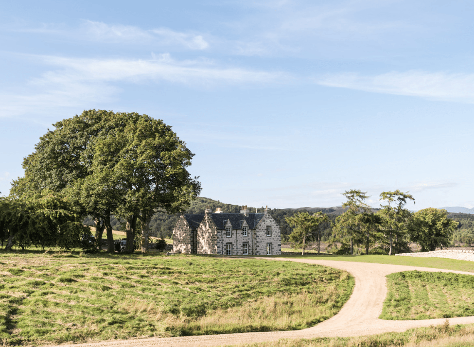 Wide view of a grassy area with trees, dirt roads and an old stone house, with mountains in the distance