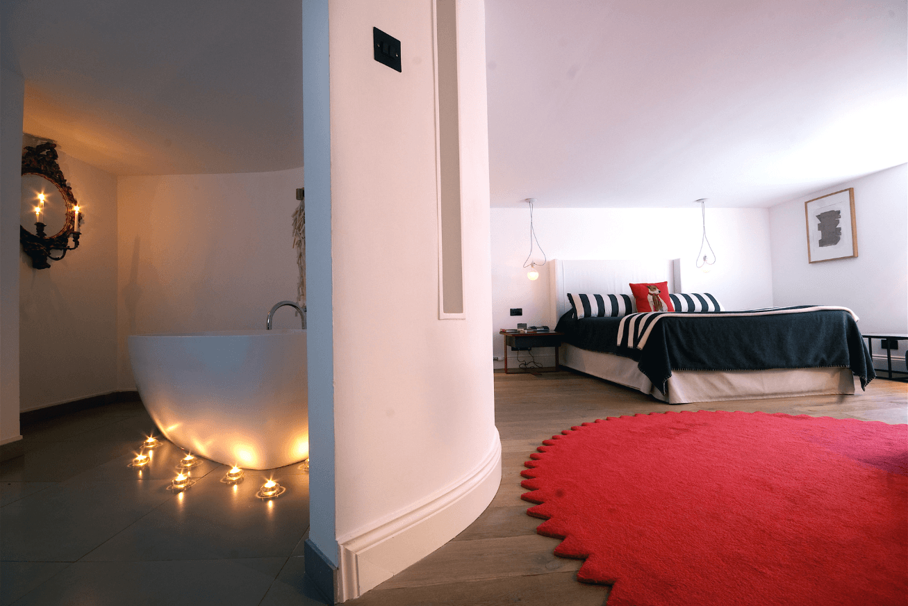 Modern hotel room with a bed, red rug and candlelit bathroom with a freestanding bathtub