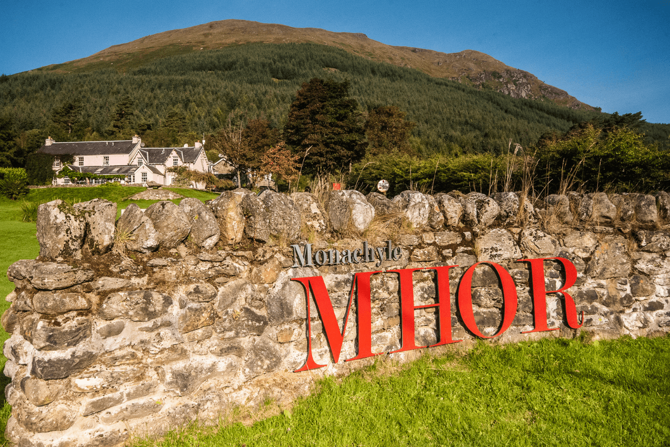 A stone wall with a sign reading 'Monachyle MHOR' and a large pink building and mountain behind it