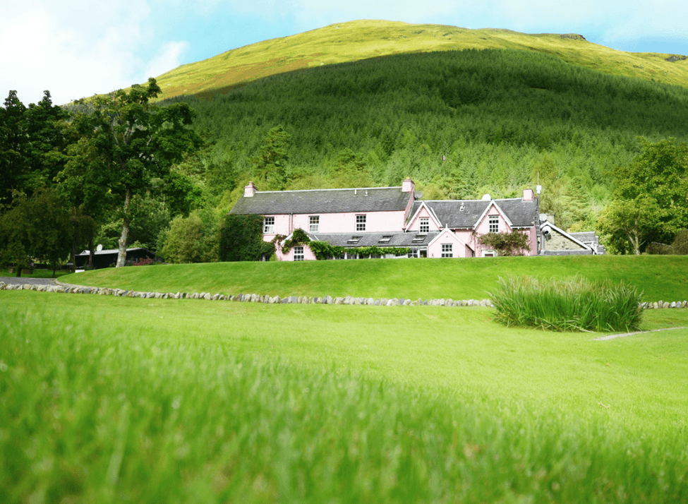 Wide view of a green grassy field with a large pink building in the background and a mountain behind it