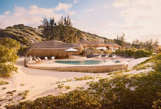 A curvy swimming pool in front of thatched-roof buildings on a sandy beach landscape under a soft sky.