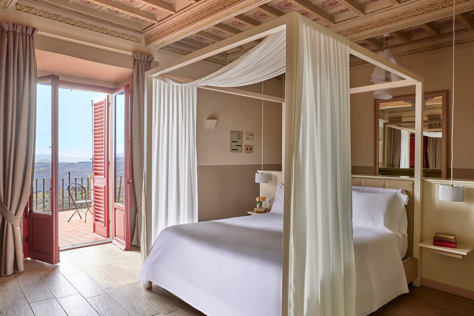 Interior of a bedroom with white bed and curtains around it, tile floors and a red door opening out onto a terrace.