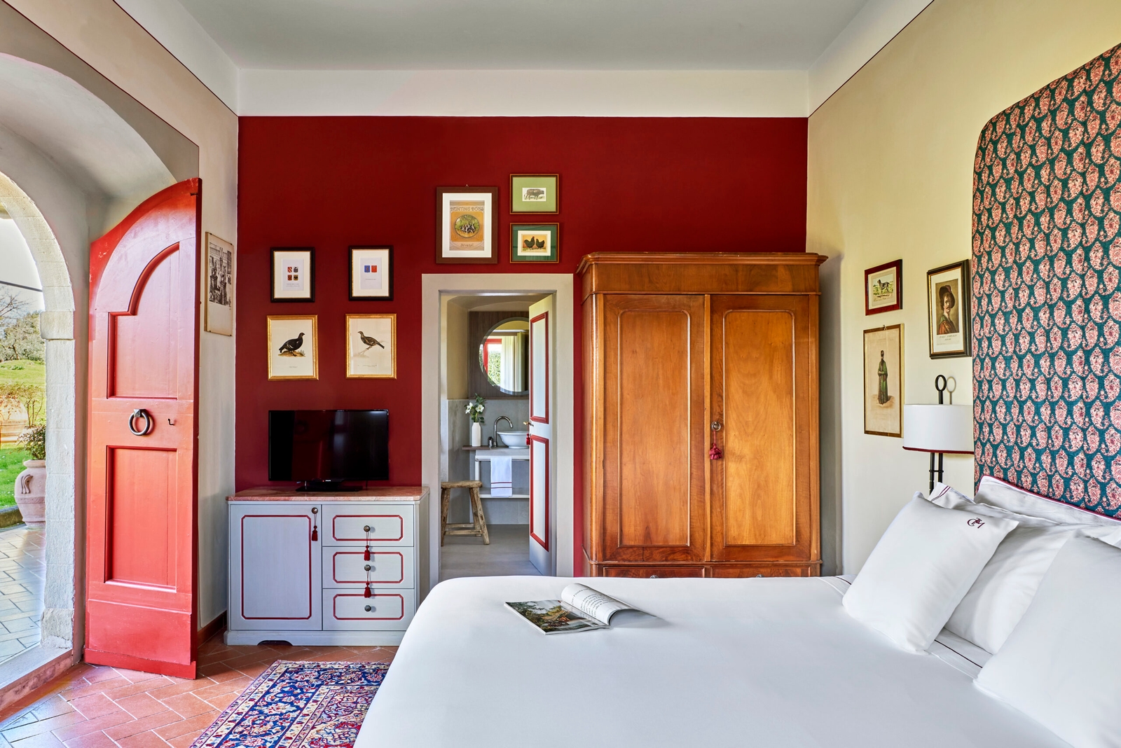Interior of a bedroom with white bed in the foreground and a wooden wardrobe and chest of drawers on either side of a door, with a red accent wall.
