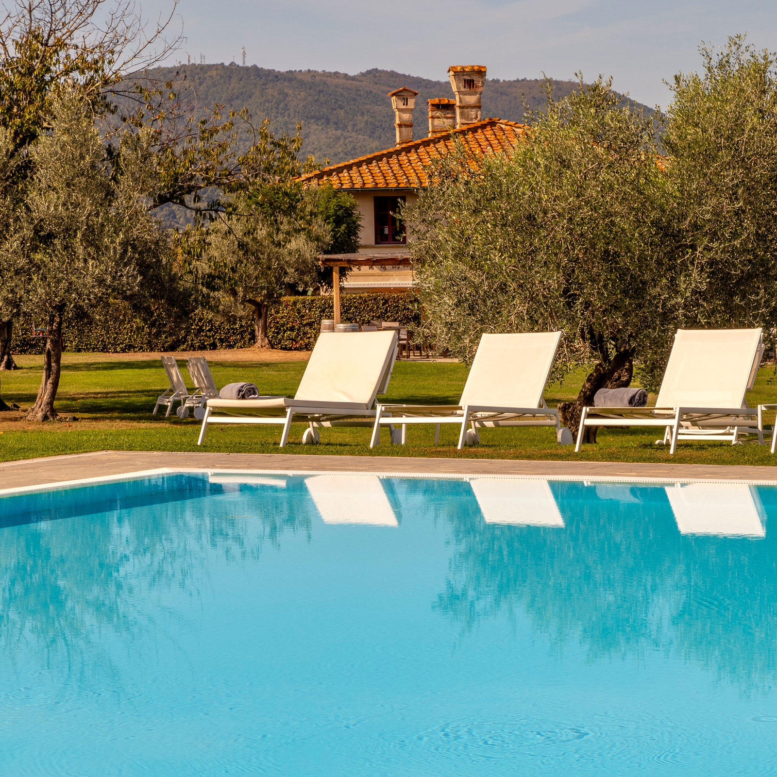 Three lounge chairs set up beside a turquoise swimming pool, with lawns, trees and the villa in the background.