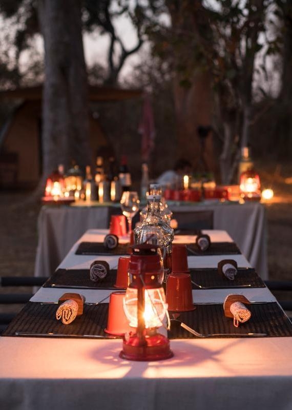 Close-up of a long safari dining table with red lanterns and wine glasses set in a wooded campsite at twilight.
