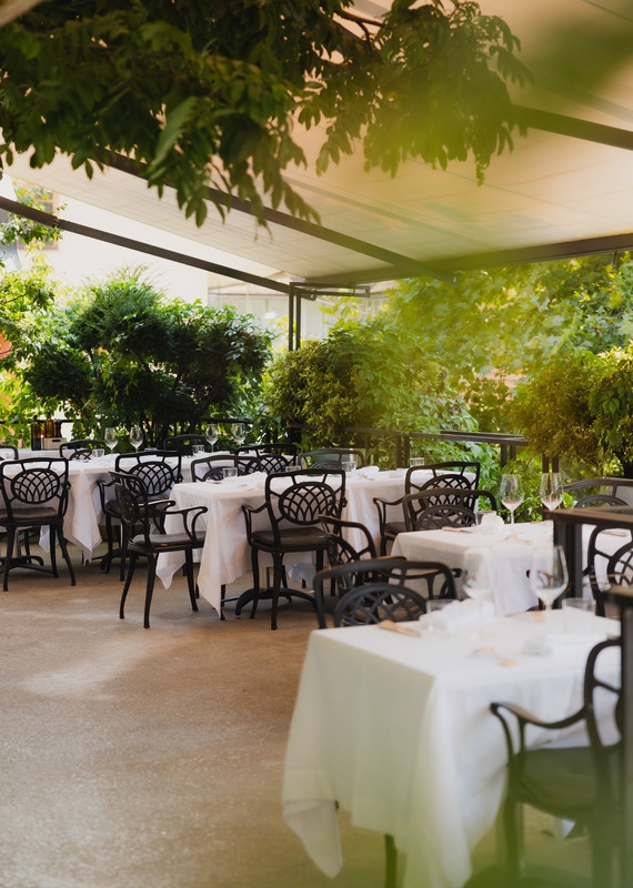 Outdoor restaurant terrace with white linen tables and black ornate chairs under a shaded garden canopy.