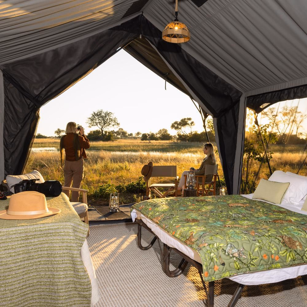 Inside a safari tent showing a bed with an animal print duvet and a person looking at the landscape with binoculars.