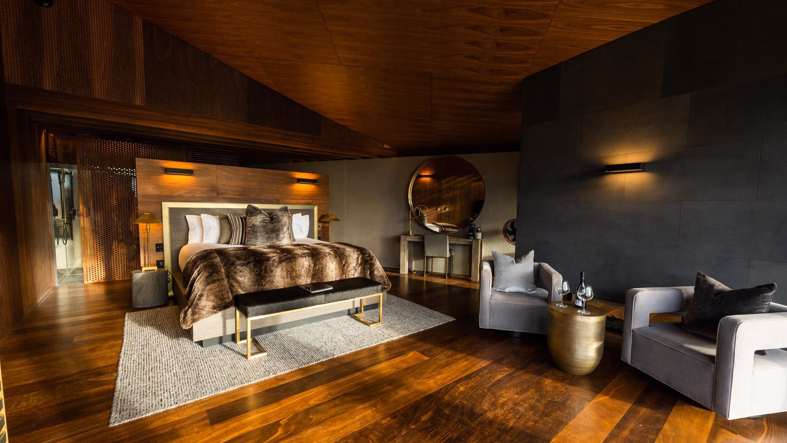 Modern luxury bedroom at Lindis Lodge with wood walls, a large bed with a fur throw, and two grey armchairs.