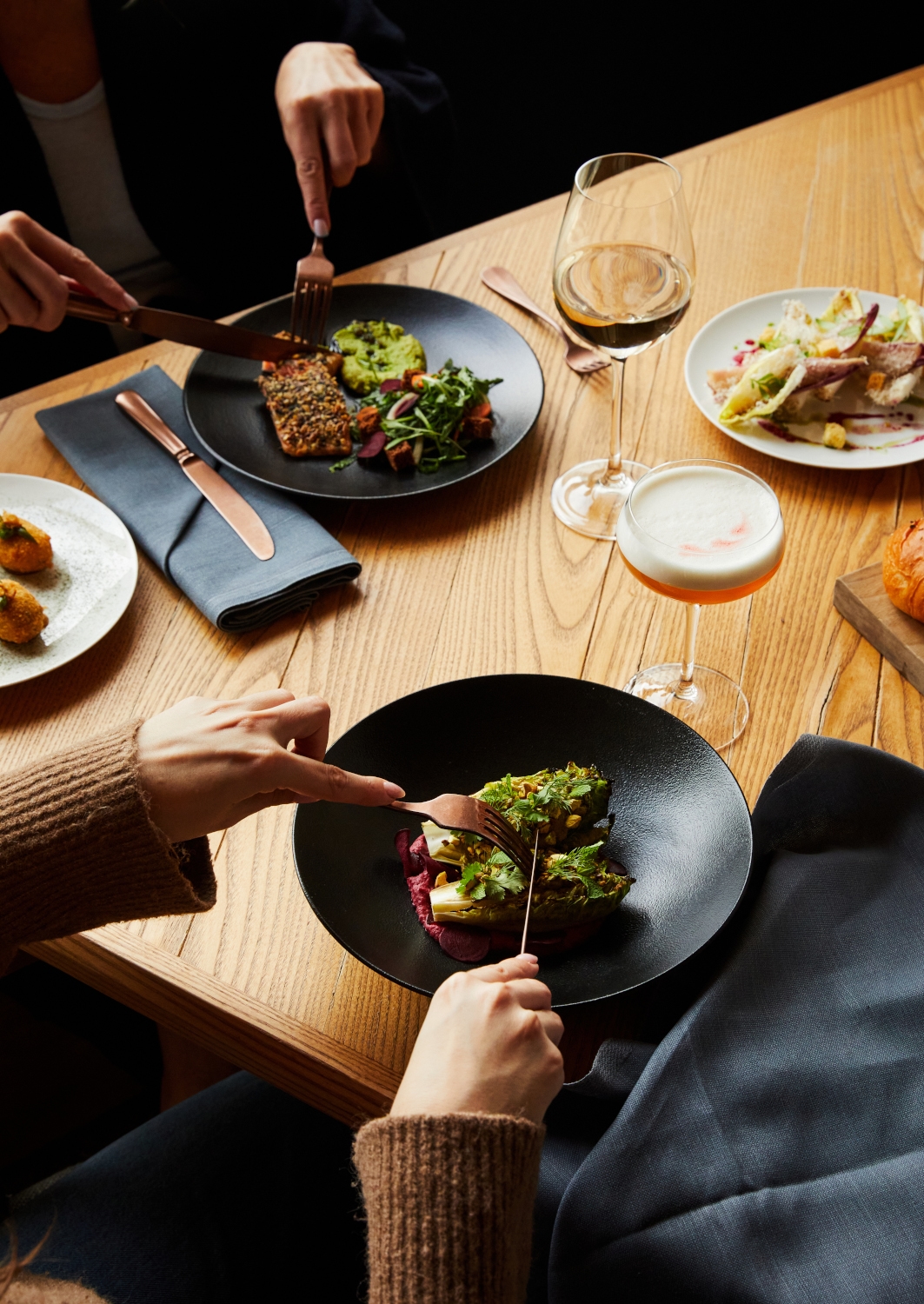 Overhead view of a light wood table with several plates of food, glasses of wine and people using forks and knives