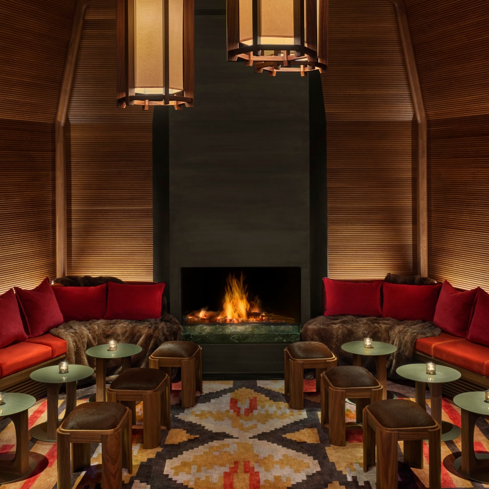 Interior of a bar lounge area with low green tables, low banquettes with red cushioins, a patterned carpet and an open fireplace in the back, with wood walls.