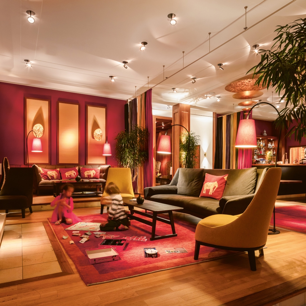Interior of a room with red walls and carpet, brown and dark grey sofas and chairs and children playing on the carpet.