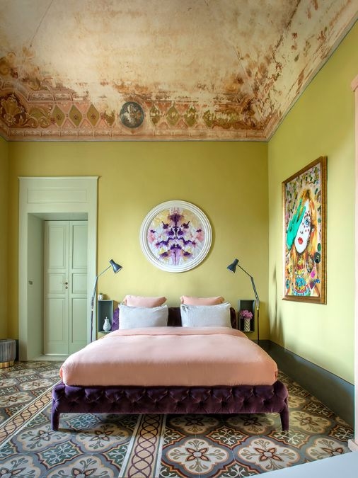 A bedroom with bright green walls and modern paintings hanging above a purple upholstered bed