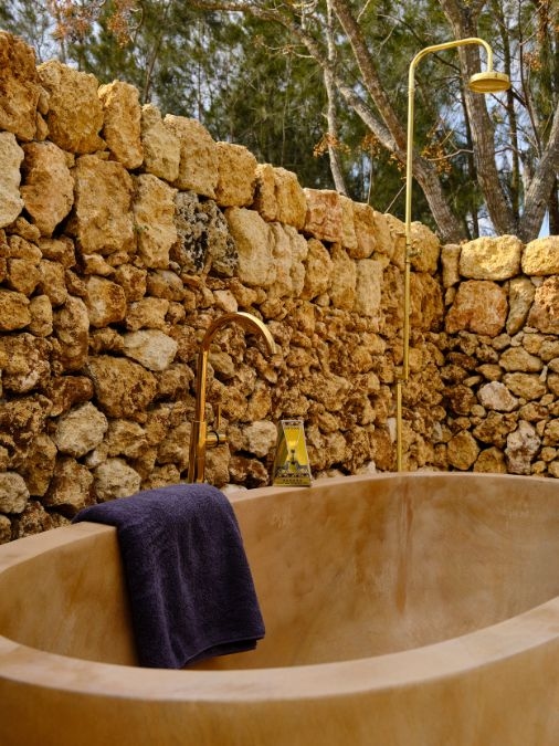 An outdoor sandstone bathtub with golden fixtures set beside a tall stone wall
