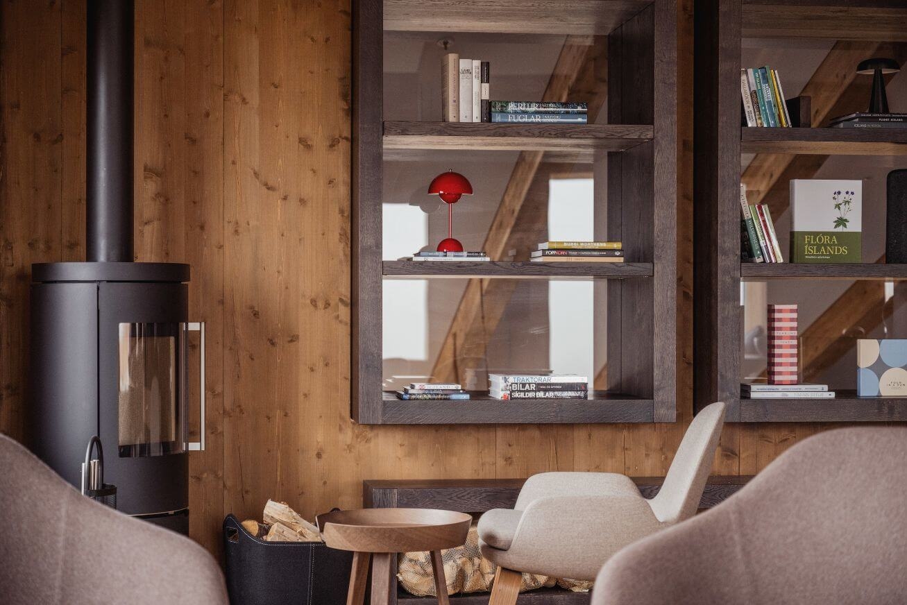 Wooden bookshelves with books and a red lamp beside a cylindrical fireplace and a chair.