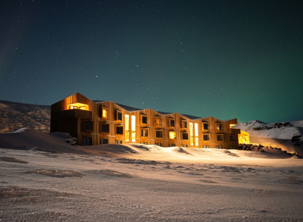 An exterior wide view of Highland Base lit up at night, with a snowy landscape around it and the glow of the northern lights.