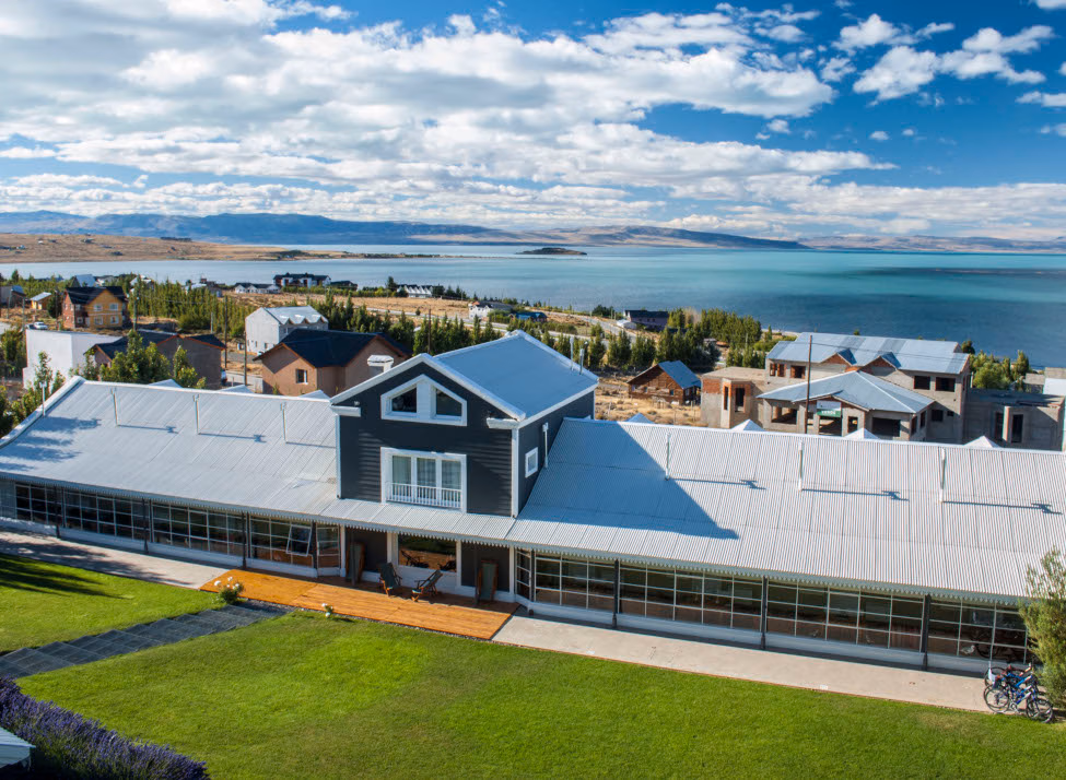 Aerial shot of the hotel with a green lawn in the foreground and scattered houses and trees in the background and the lake and shoreline beyond.
