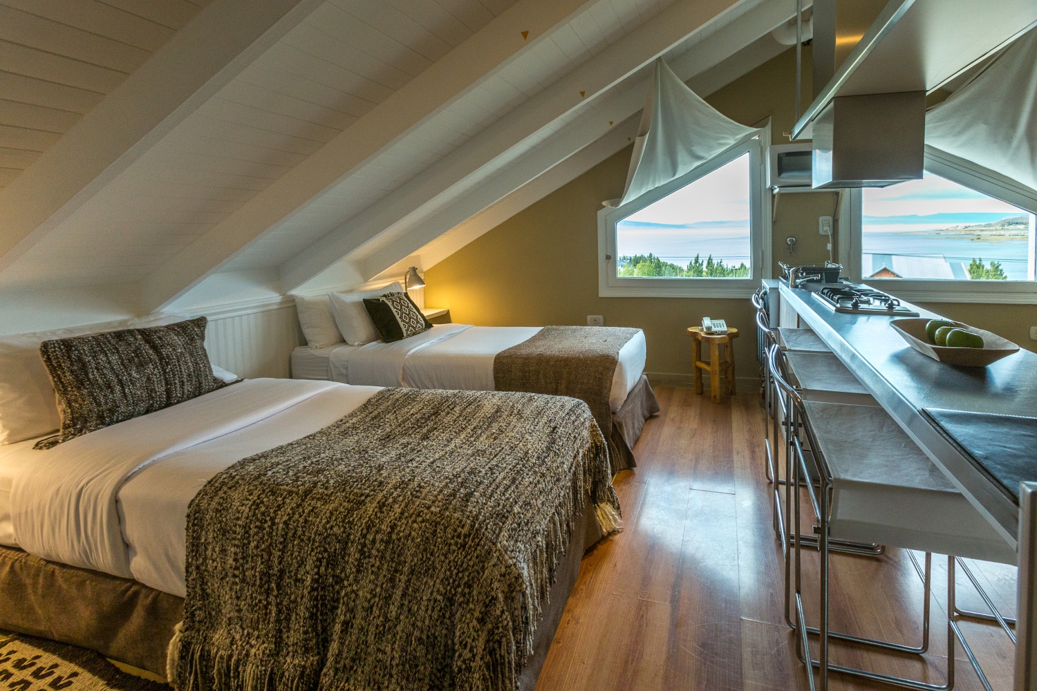Bedroom with two double beds dressed in muted colours, wood floors and a slanted ceiling, with a small window and lake views.