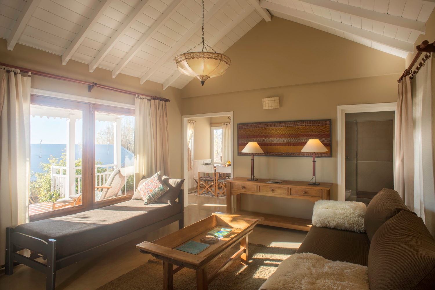 Living room area with big windows overlooking the lake. Inside is a brown sofa with white throw pillows, a side desk with two lamps, a chaise lounge underneath the windows and a coffee table in the centre.