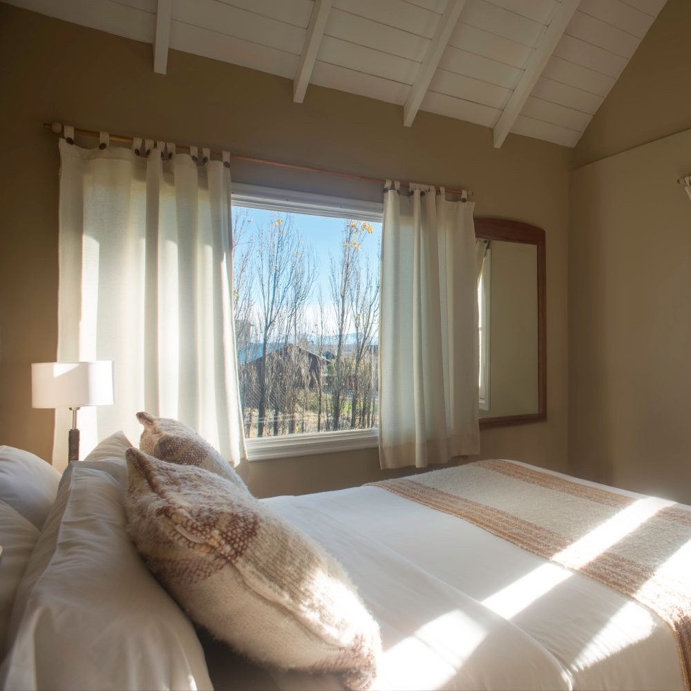 Double room dressed in white and beige bedding with a window and white curtains, and white timber ceiling.