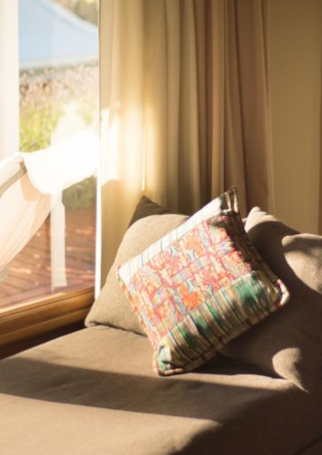 A beige chaise lounge with colourful woven pillow beside a brightly lit window.