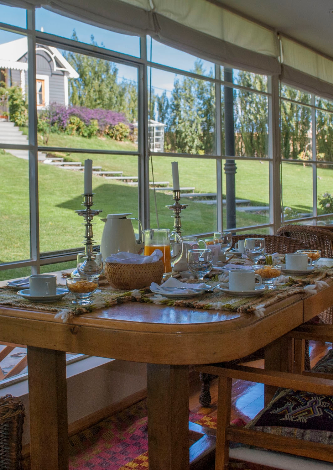 Wooden table set for breakfast with juices, coffees and plates, beside big windows looking out onto a green lawn.