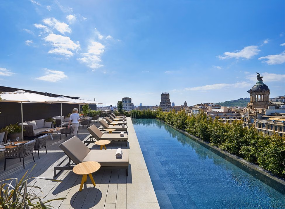 A long and narrow rooftop pool with sun loungers, a dining area and views of Barcelona