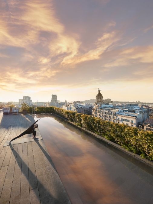 A long and narrow rooftop pool with a woman doing yoga beside it and sunset views of Barcelona