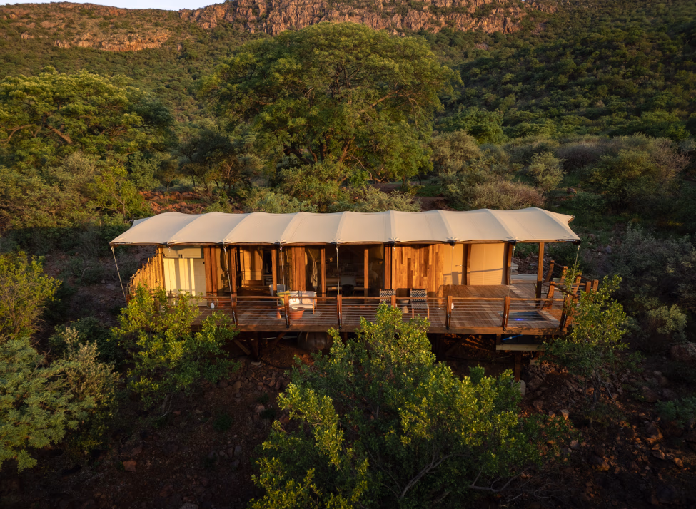 Exterior shot of a cliffside tented suite with canvas roof and wraparound wooden deck set among green trees with mountains in the distance.