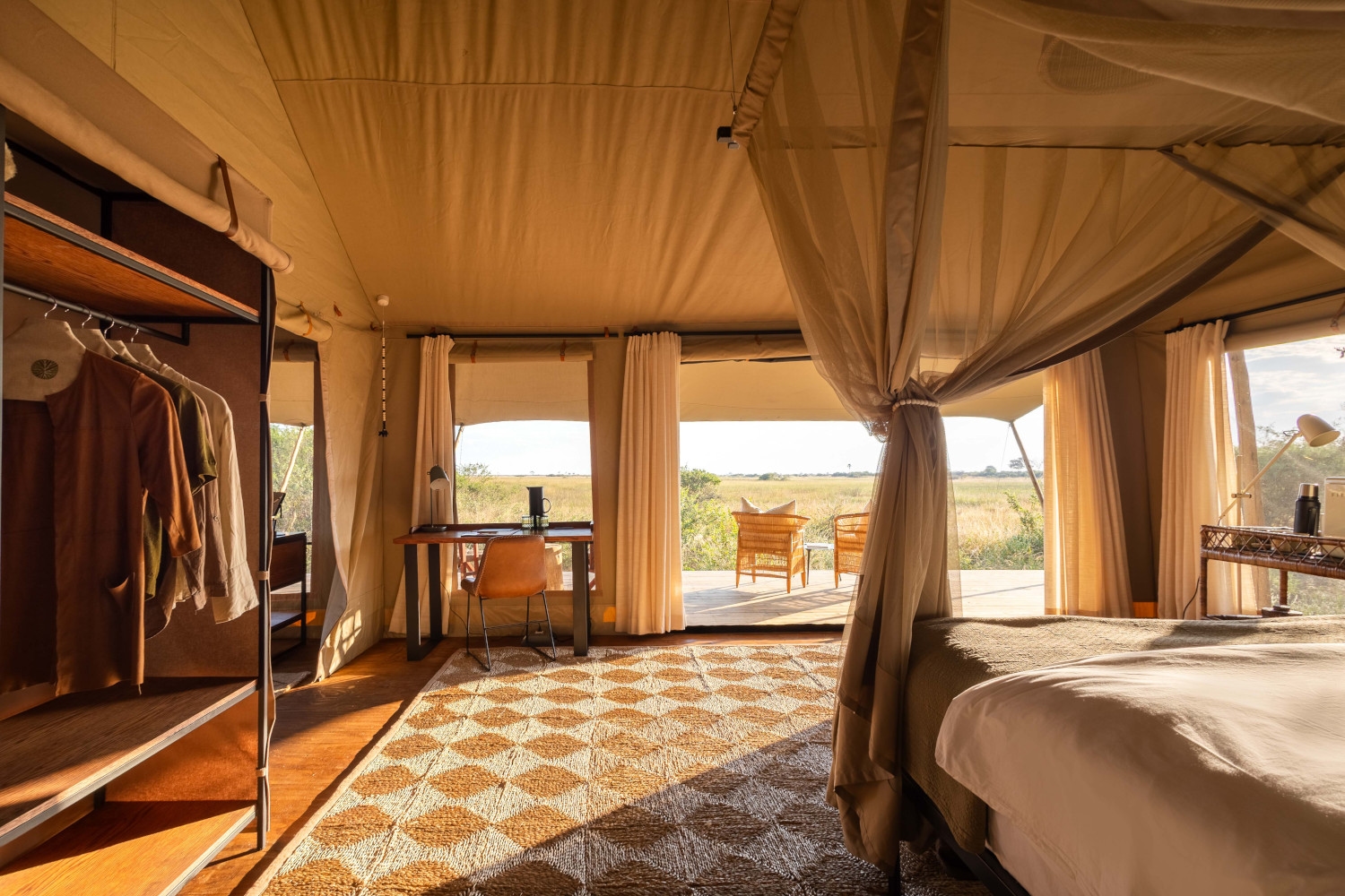 Interior of a safari tent bedroom with a bed on the right and gathered mosquito net and open windows looking into a deck with two chairs and the bush beyond.
