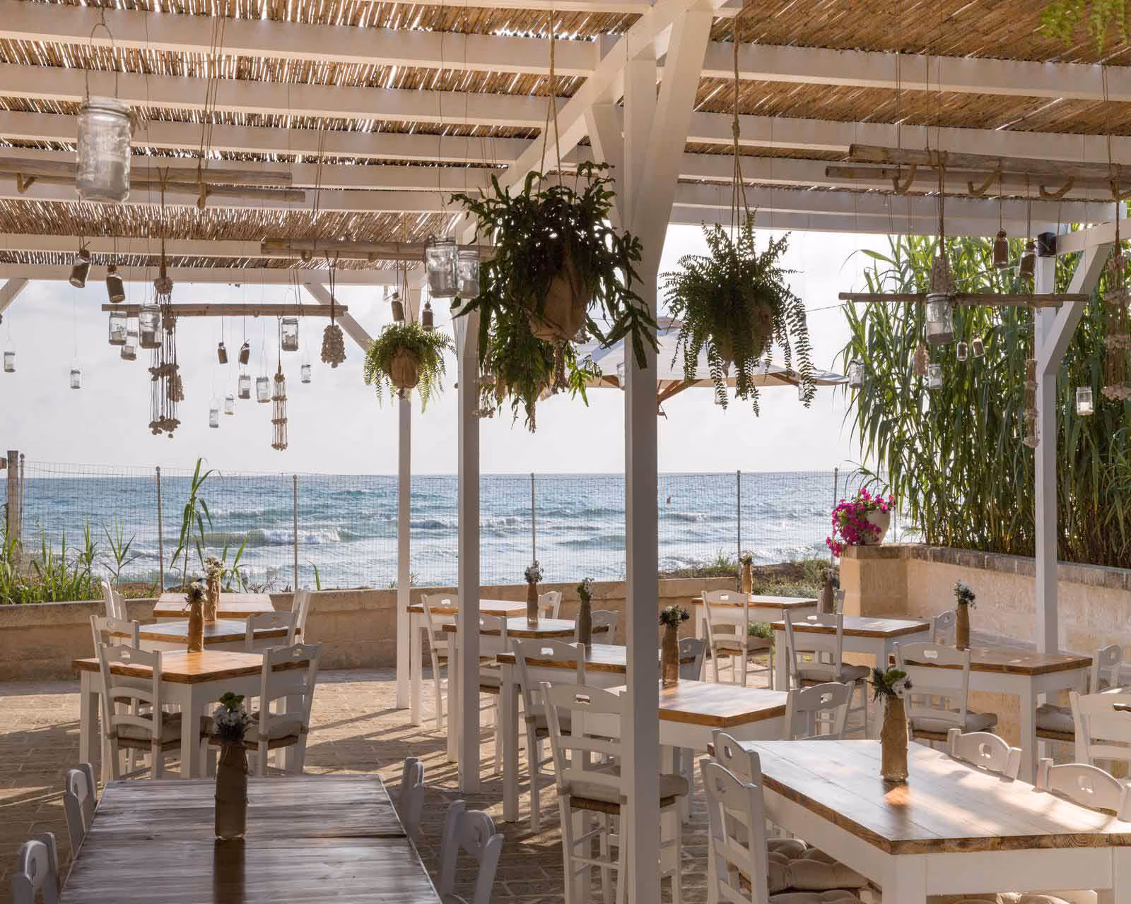 An open-air restairant with white and natural wood tables and chairs, a thatched bamboo roof and open views of the sea. Potted plants and candles in jars hang from the ceiling.