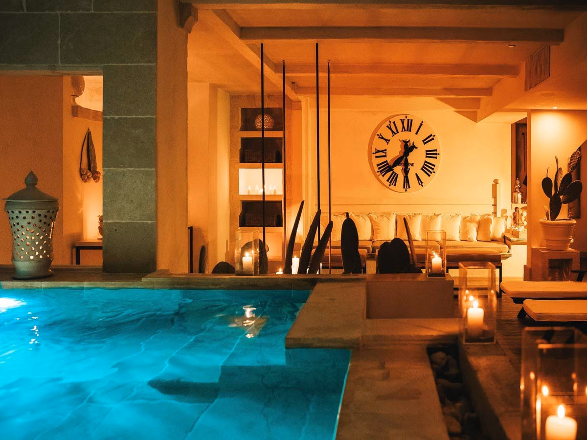 A spa setting with a thalassotherapy pool that has deep steps, in a candlelit space with lanterns, a long sofa with cushions and a big clock on the wall. The sauna is on the left behind the pool.